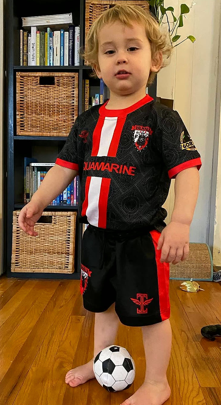 A young Valhalla in the black and red kit with the crest on his chest and the ball at his feet